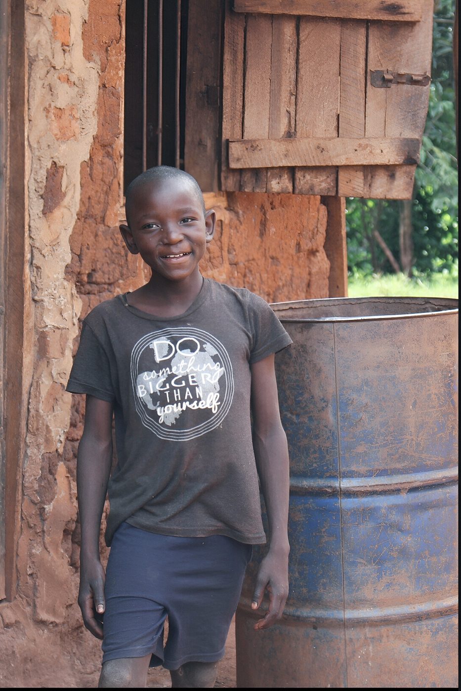A young boy from the Ekubo community in Uganda smiling outside his home, wearing a shirt that reads 'Do Something Bigger Than Yourself'
